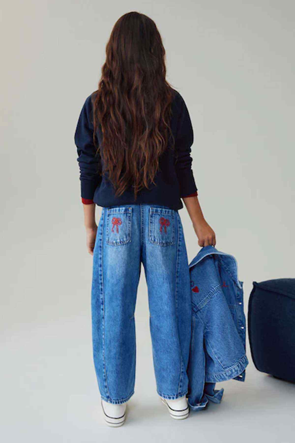 Kids blue denim jeans with red embroidered motifs, back view holding denim jacket