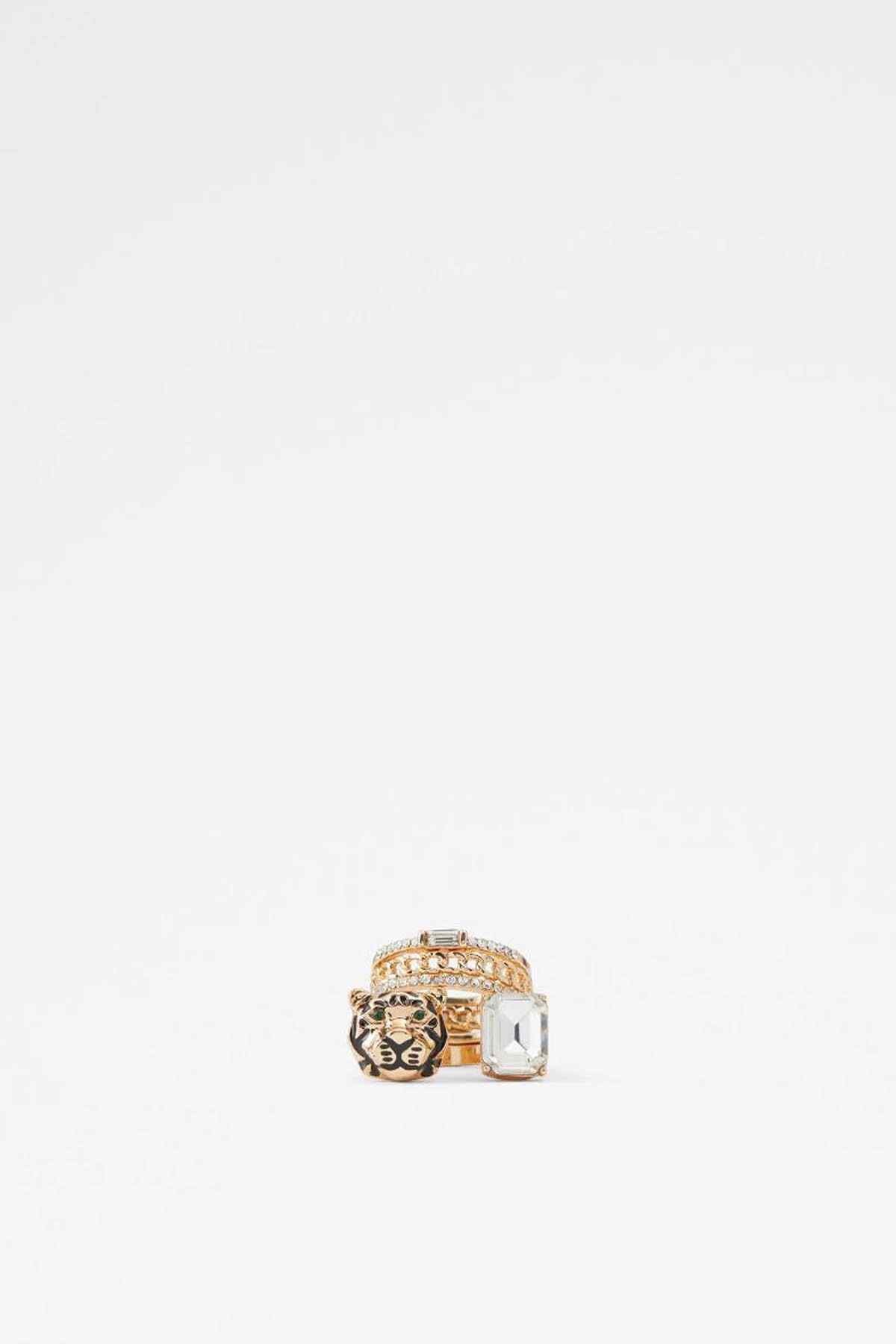 Gold-tone ring with stacked bands, clear square stone, and lion head charm.
