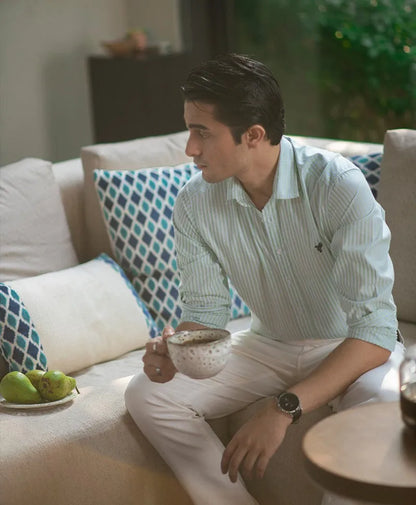 Men's striped button-up shirt with white pants and belt, seated with wristwatch