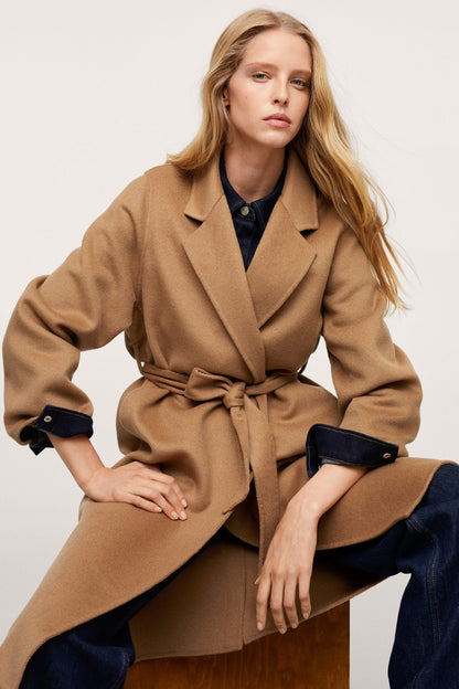 Tan belted long coat with dark cuffs, lapels, and tied waist belt detail