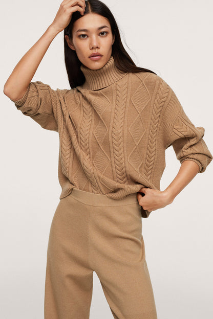Tan cable-knit turtleneck sweater with long sleeves, paired with trousers