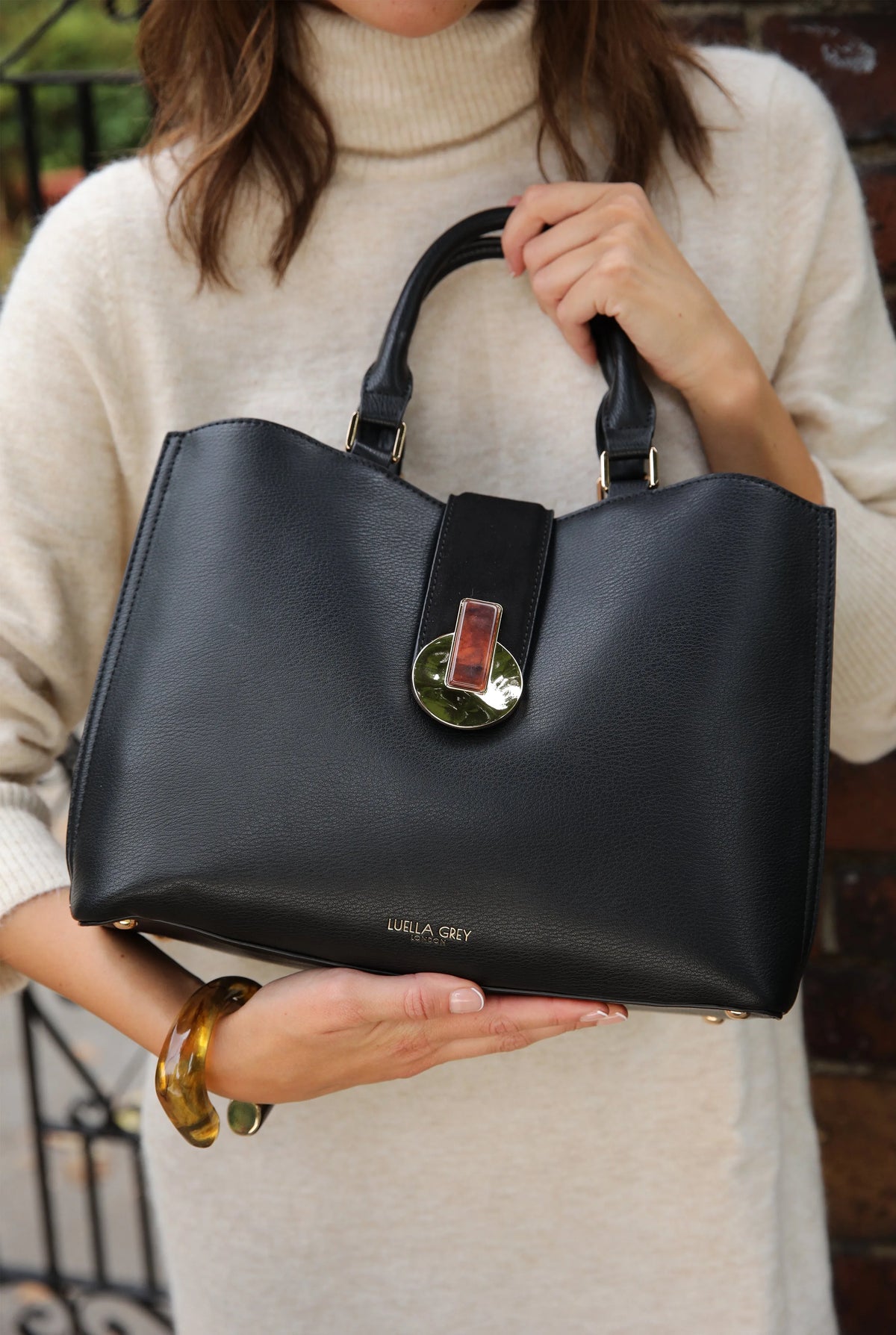 Black tote bag on person with top handles, center clasp, and gold-tone hardware