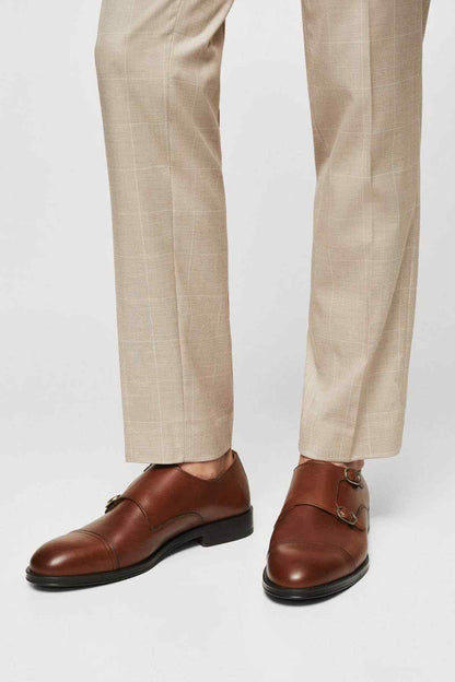 Brown leather double-buckle loafers on feet with light plaid trousers