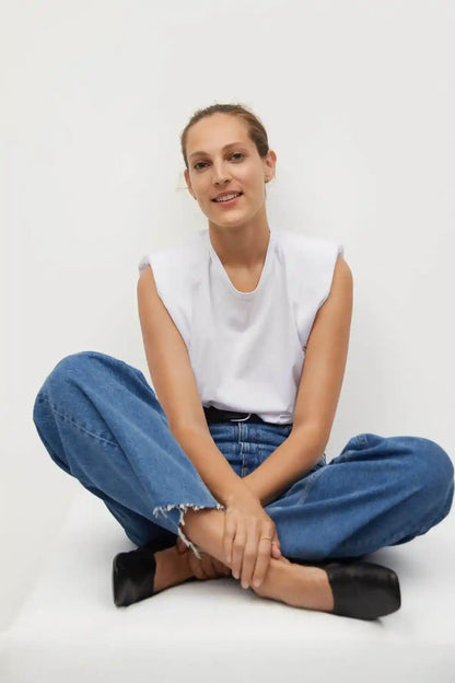 White sleeveless top with blue jeans and black belt, seated with legs folded