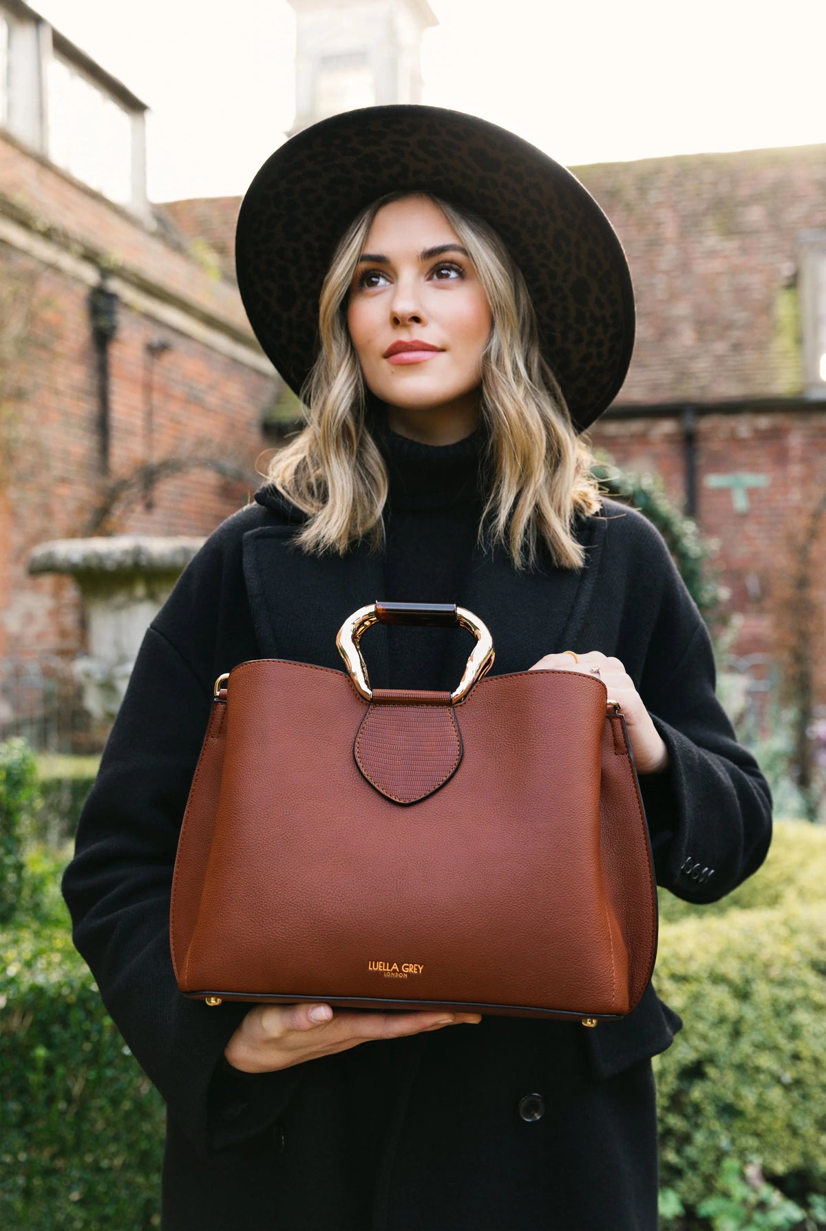 Brown handbag held in hands with top metal clasp and short front logo