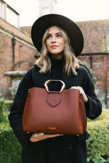 Brown handbag held in hands with top metal clasp and short front logo