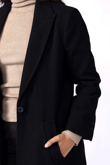 Black long coat detail near lapel and front pocket; beige turtleneck visible.