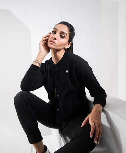 Black fitted button-front jacket with long sleeves and chest logo, model seated in a side view